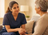 Nurse demonstrating empathy during an OET Speaking role-play while sitting on a sofa in a warm, welcoming environment, wearing fitted navy blue scrubs.