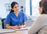 International nurse in blue scrubs practising an OET Speaking role play with a patient in a UK clinical consultation room.