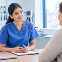 International nurse in blue scrubs practising an OET Speaking role play with a patient in a UK clinical consultation room.