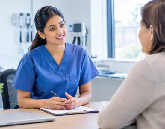 International nurse in blue scrubs practising an OET Speaking role play with a patient in a UK clinical consultation room.