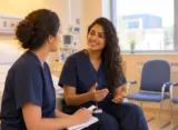 Two nurses in blue scrubs having a warm discussion in a hospital patient room near the window.